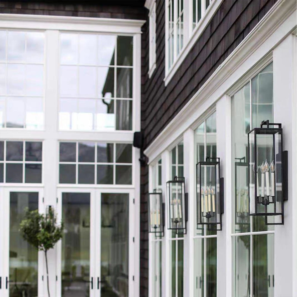Black forged iron clear glass outdoor wall lantern sconce mounted on a white exterior patio wall with black framed doors and windows.