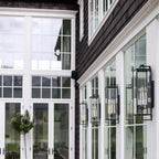 Black forged iron clear glass outdoor wall lantern sconce mounted on a white exterior patio wall with black framed doors and windows.