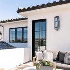 Black forged iron clear glass outdoor wall lantern sconce mounted on a white exterior patio wall with black framed doors and windows.