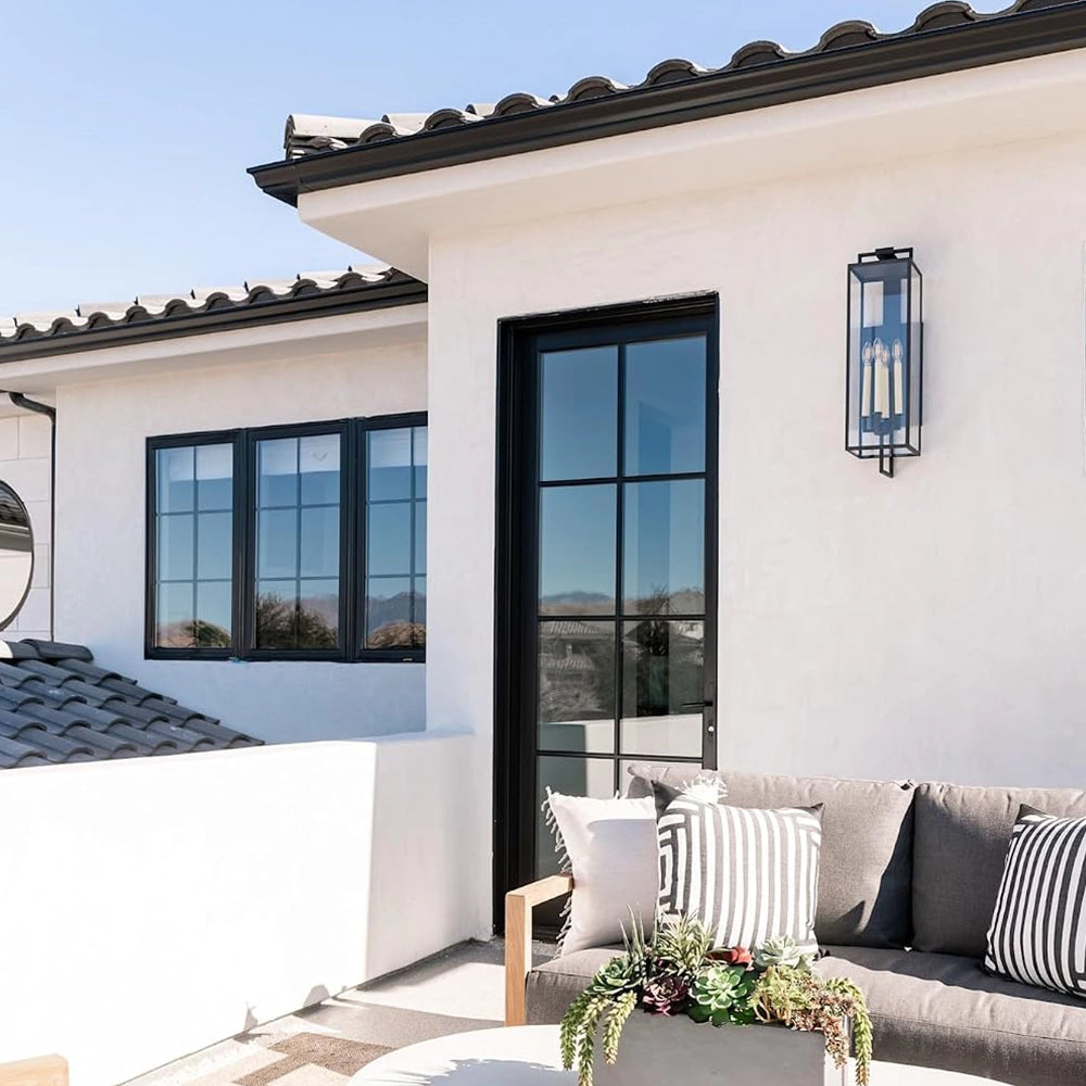Black forged iron clear glass outdoor wall lantern sconce mounted on a white exterior patio wall with black framed doors and windows.